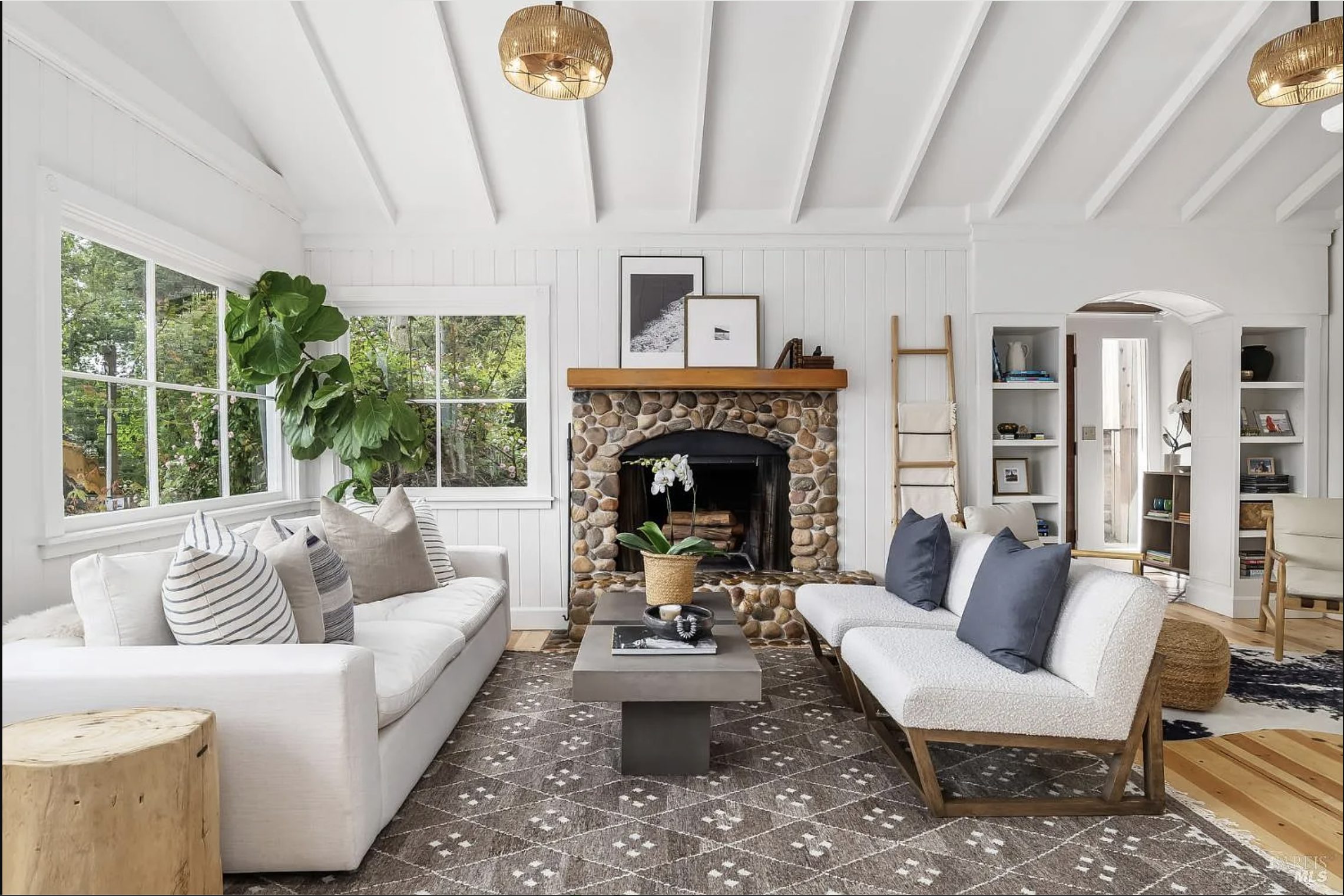 A bright and inviting living room featuring exposed beam ceilings, a handcrafted river rock fireplace, oversized picture windows, natural light, warm wood accents, and curated organic-modern furnishings that reflect the timeless character of a 1950s Craftsman-inspired Marin County home.