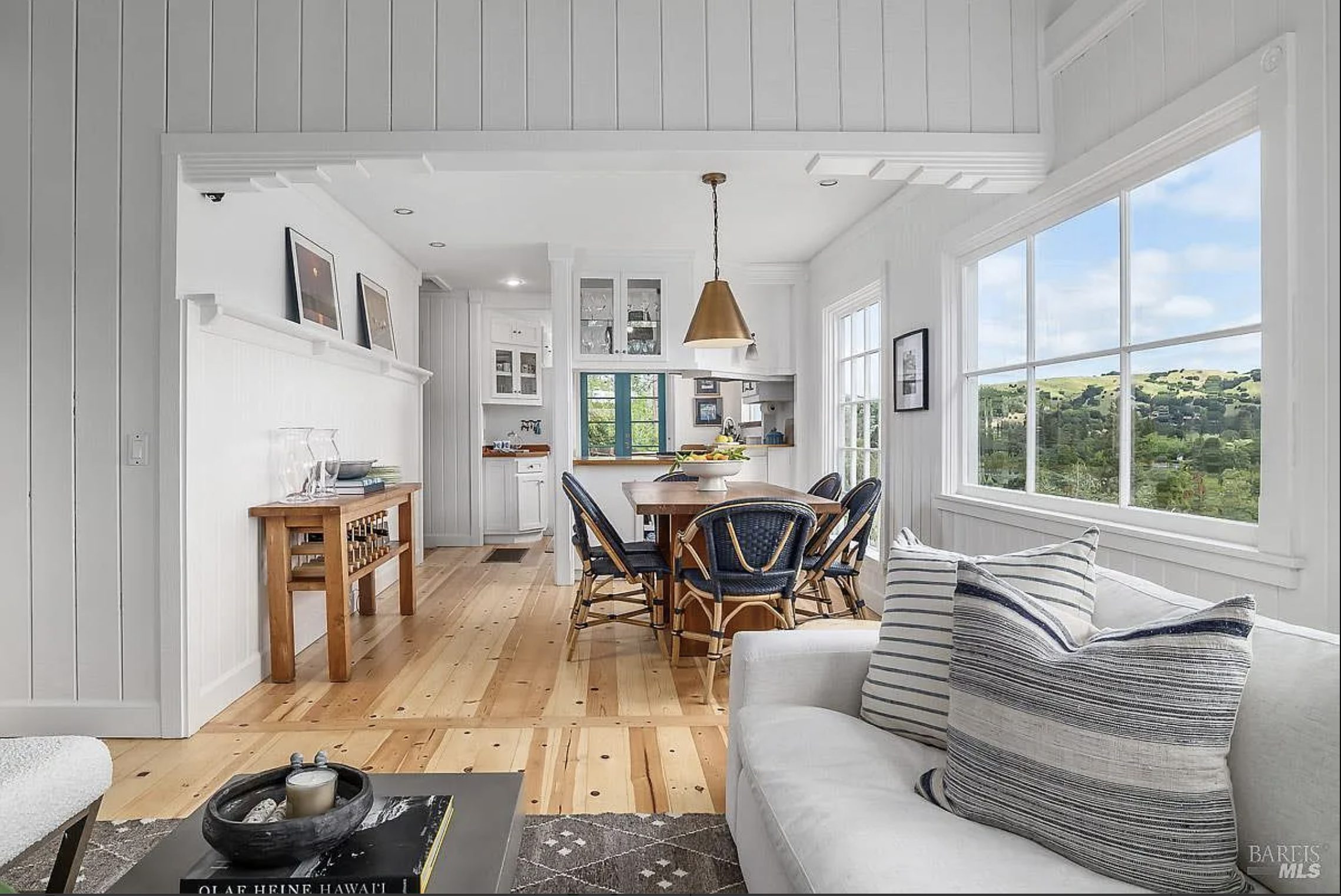 An open-concept dining and living space with original wood flooring, vaulted ceilings, panoramic hillside views, abundant natural light, and seamless flow between the dining area and kitchen—showcasing the warmth and simplicity of mid-century Marin County architecture.