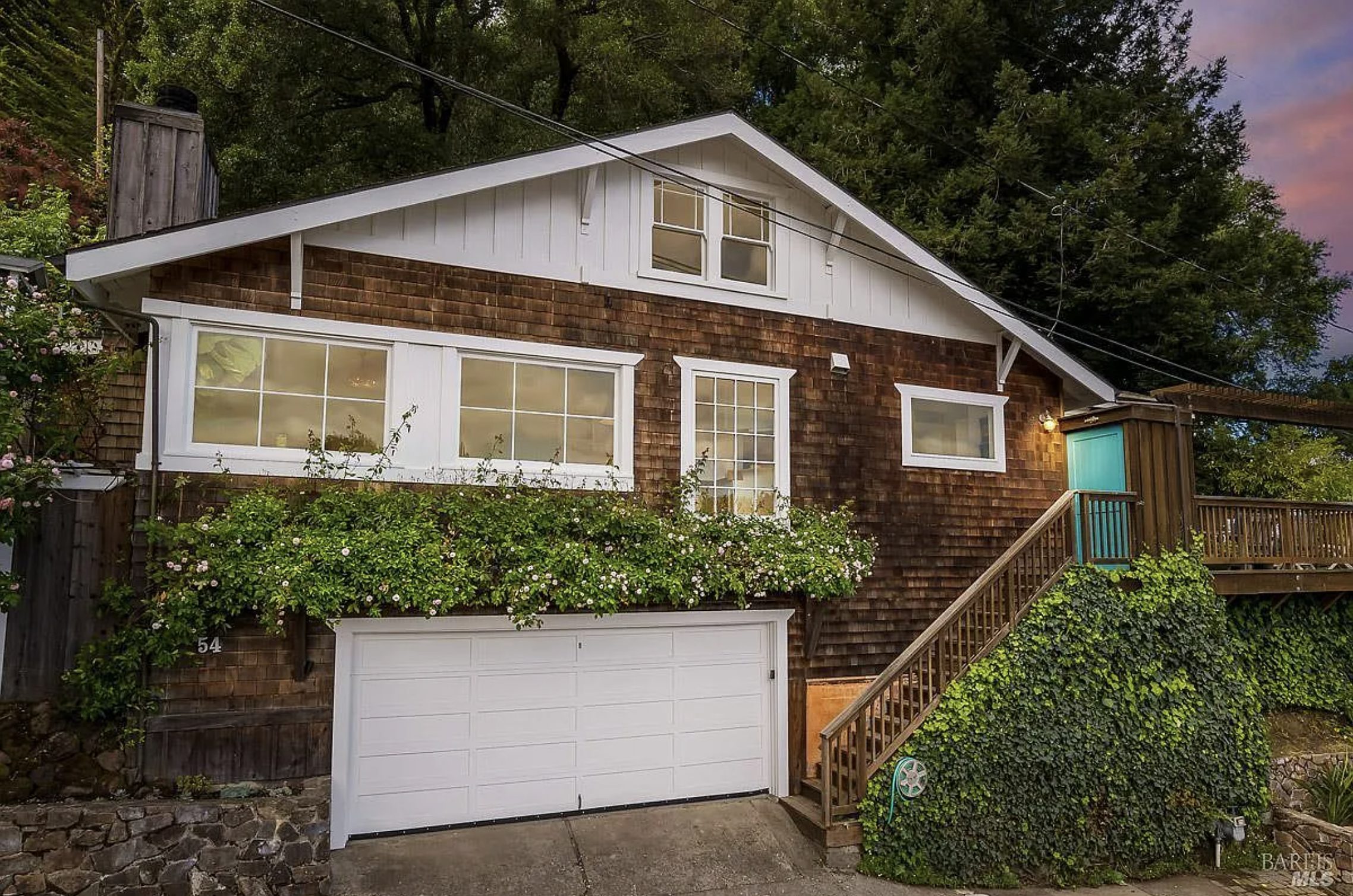 The exterior of a classic 1950s Craftsman-inspired home in San Anselmo featuring cedar shingle siding, clean mid-century rooflines, lush mature landscaping, ivy-covered retaining walls, warm exterior lighting, and a private hillside setting surrounded by towering Marin County trees.